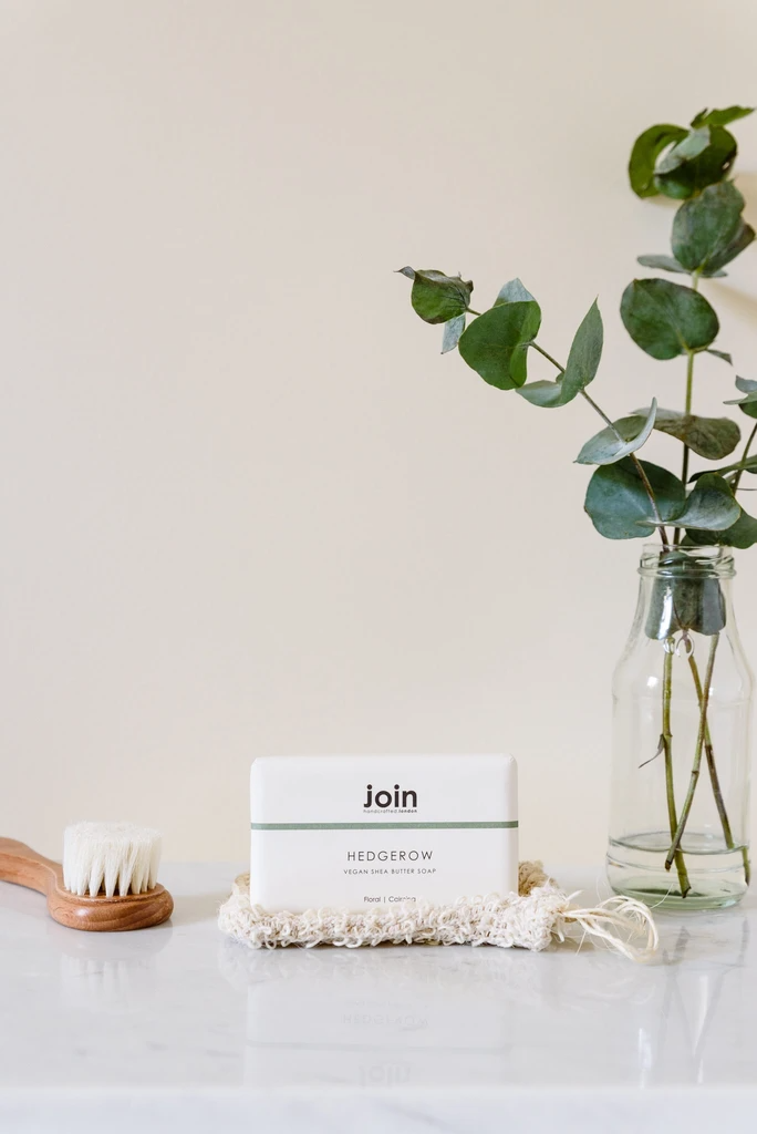 A packaged bar of Hedgerow soap with a floral design in a minimalist recycled wrapping, placed on a marble surface with a vase of green leaves in the background.