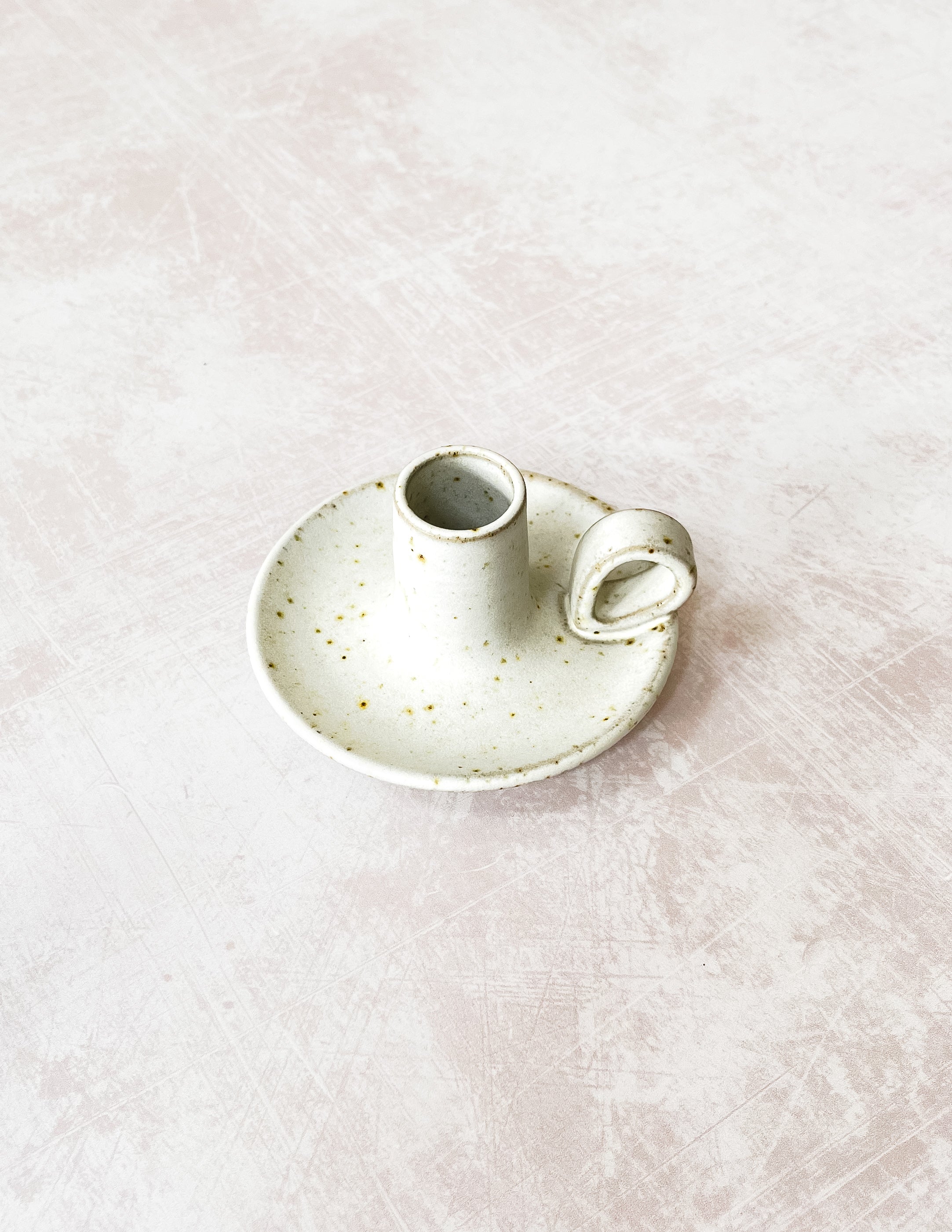 A handmade stoneware candlestick holder with a loop handle on a textured surface.