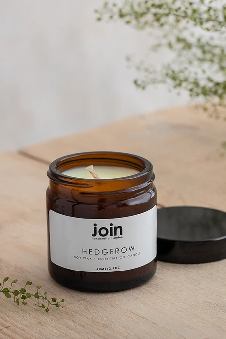 A brown glass jar candle with the label 'JOIN' and 'HEDGEROW' is displayed on a wooden surface with a beige pot and green foliage in the background.
