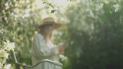 Woman in whimsical dress and straw hat under blossom tree next to her bike on her phone