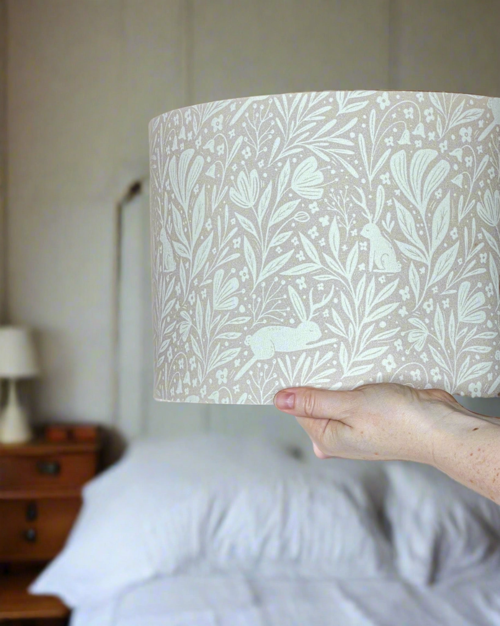 Whispering Woodlands drum lampshade with enchanting hares among woodland flowers in white, set against a light delicate pink background.