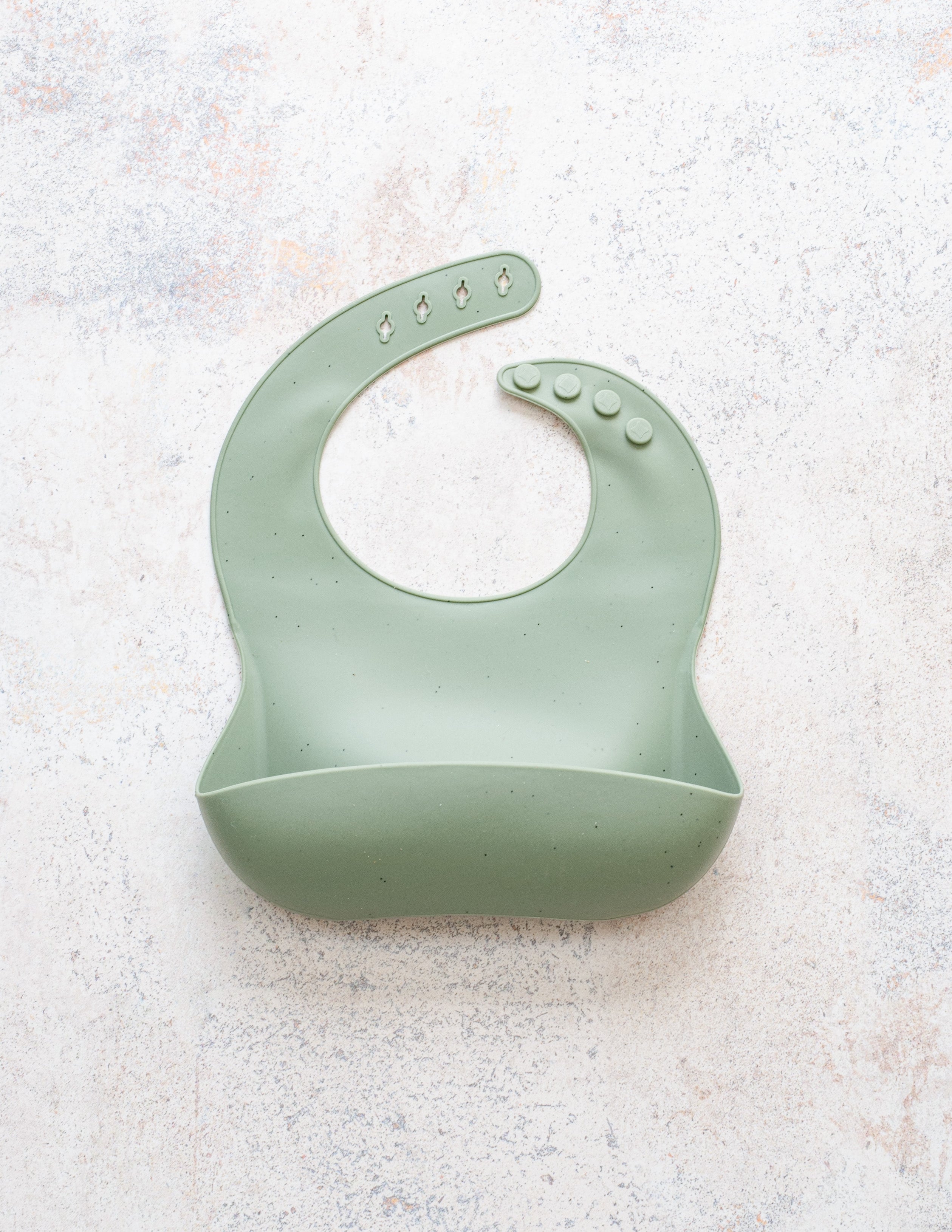 A green silicone bib with a deep front pocket and rounded neck fasteners, displayed against a speckled grey background.