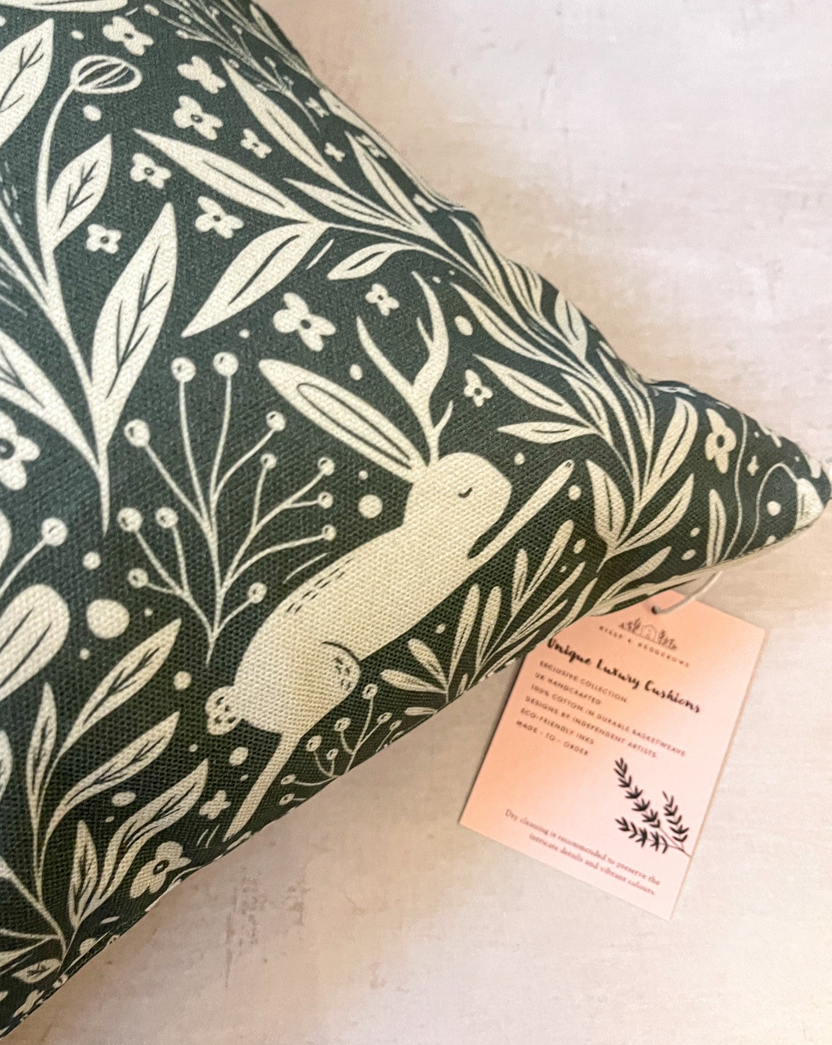 Whispering Woodland Cushion featuring an enchanting botanical pattern with a leaping hare with one ear and one antler, set against a deep blue-green background with a murky cream foreground.