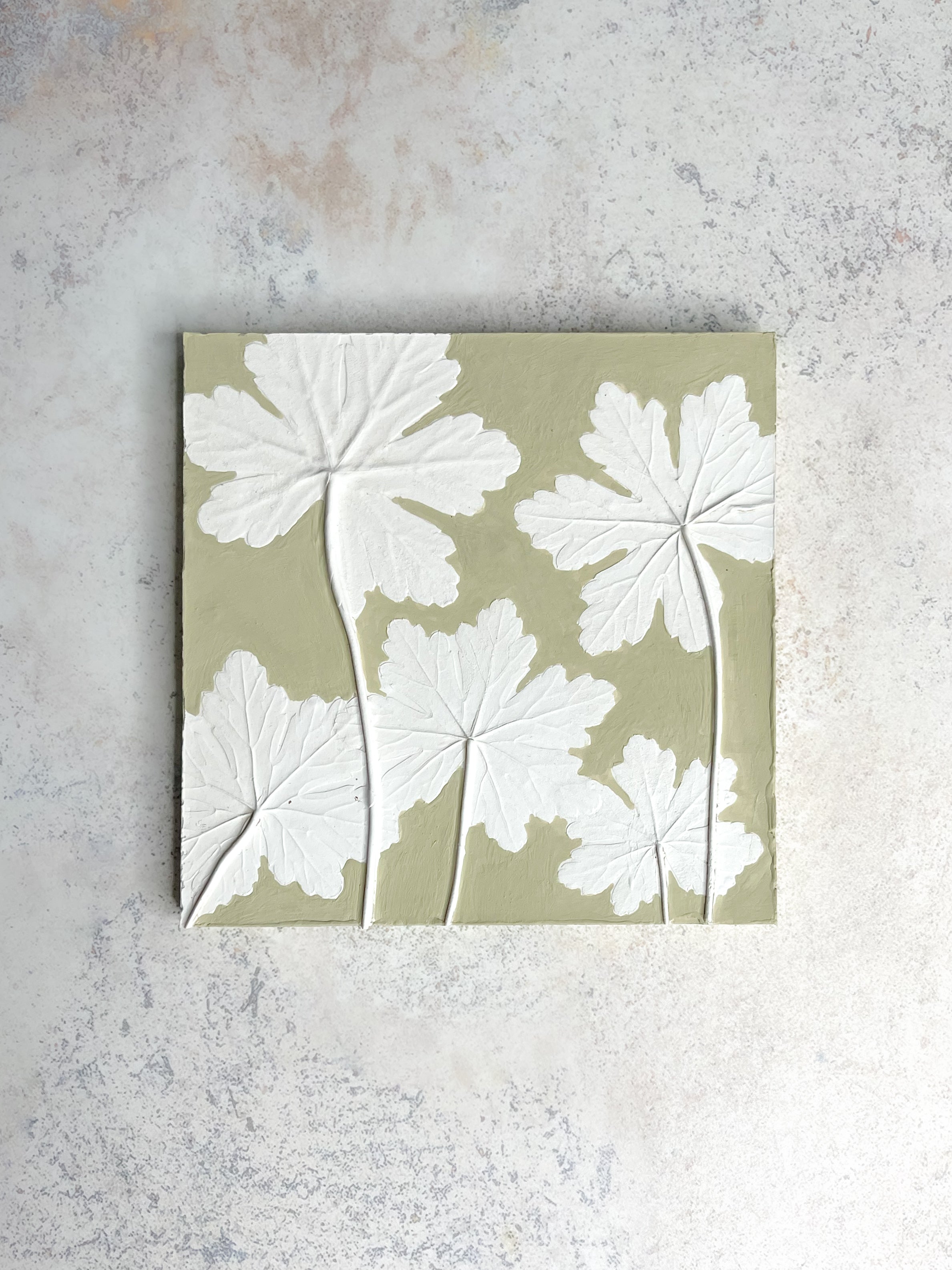 Handcrafted botanical relief tile featuring geranium leaves, hand-painted in soft mossy green, symbolising health and positive energy.